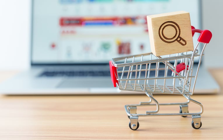 Photo of a shopping cart with a magnifying glass icon block and laptop computer with marketplace website